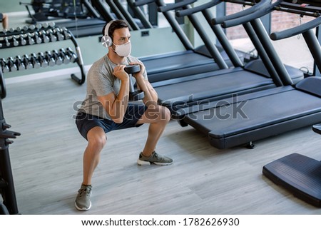 Athletic man using dumbbell while exercising squats and wearing protective face mask in a gym. 