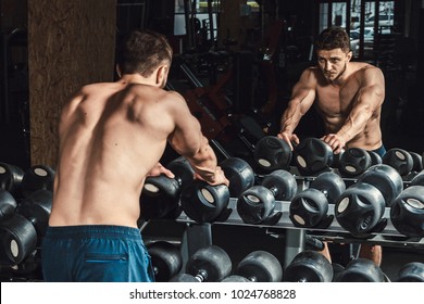 athletic man waiting and looking at himself before lifting dumbbells in front of the mirror