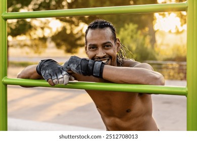 Athletic man wearing training gloves resting on a green bar in an outdoor park. The sun sets behind the trees, creating a warm atmosphere.  - Powered by Shutterstock