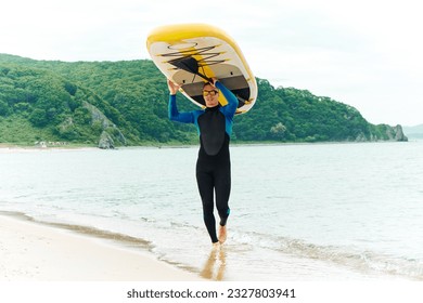 An athletic man in a wetsuit carries a SUP board on his head along the sandy seashore. Extreme recreation.