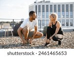 Athletic man in white t-shirt and woman in black top tying shoelaces before exercising on roof terrace. Cheerful couple preparing for running at summer morning against background of cityscape.