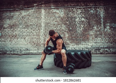 Athletic man working out with a dumbbell in front of brick wall while sit on big tire. Strength and motivation. Outdoor workout. Exercise for the muscles biceps