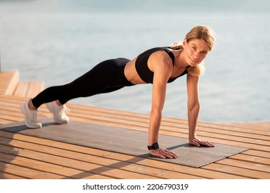 Athletic Middle Aged Woman Making Straight Arm Plank Exercise While Training On Fitness Mat Outdoors, Smiling Sporty Female In Activewear Exercising On Wooden Pier Near Water, Copy Space - Powered by Shutterstock