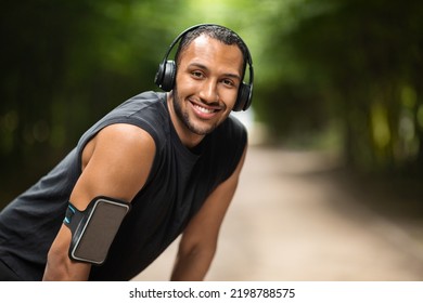 Athletic millennial african american man in black sportswear exercising in public park, using wireless headphones, armband for smartphone, listening to music while jogging, panorama with copy space - Powered by Shutterstock