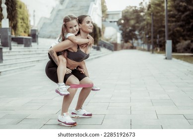 Athletic mom exercising with her little daughter on her back outdoors. Squat with fitness rubber bands. Healthy lifestyle, fitness active family concept. Training together