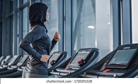 Athletic Muslim Sports Woman Wearing Hijab and Sportswear Running on Treadmill. Energetic Fit Female Athlete Training in Gym Alone. Urban Business District Window View. Side View Portrait - Powered by Shutterstock