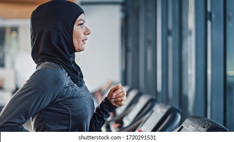 Athletic Muslim Sports Woman Wearing Hijab And Sportswear Running On Treadmill. Energetic Fit Female Athlete Training In Gym Alone. Urban Business District Window View. Side View Portrait