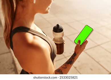 An athletic person dedicated to fitness takes a break after an intense workout, holding a smartphone with a bright green screen and a bottle of water to stay hydrated and refreshed - Powered by Shutterstock