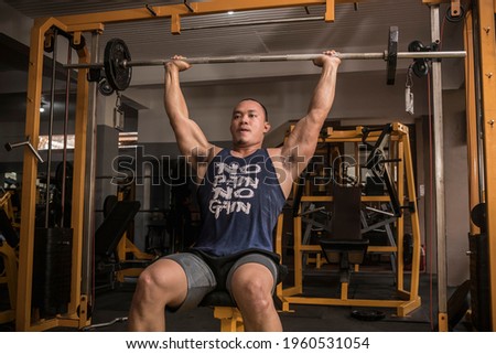 An athletic and rugged asian man does Seated Military Presses. Shoulder workout at the gym.
