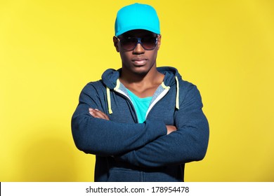 Athletic runner with sunglasses wearing blue sportswear fashion. Black man. Blue cap and sweater. Intense colors. Studio shot against yellow background.