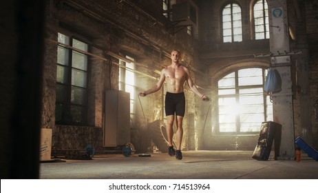 Athletic Shirtless Fit Man Exercises With Jump / Skipping Rope In A Deserted Factory Hardcore Gym. He's Covered In Sweat From His Intense Fitness Training.