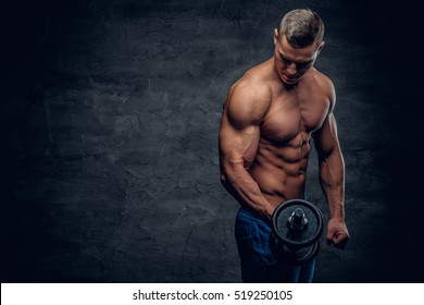 Athletic shirtless young male fitness model holds the dumbbell with light isolated on dark background.