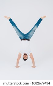 Athletic smiling barefoot young man in jeans performing a handstand with wide open legs over a white studio background
