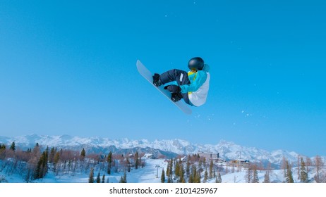Athletic snowboarder takes off into the air and performs a spinning grab trick while enjoying a sunny winter day in sunny Vogel. Active male tourist does a snowboarding trick during winter vacation.
