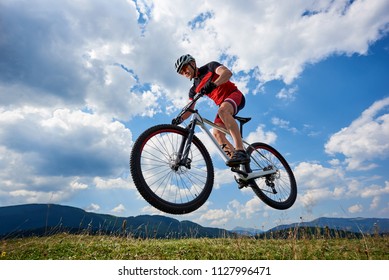 Athletic Sportsman Cyclist In Professional Sportswear And Helmet Flying In Air On His Bicycle On Summer Blue Sky With White Clouds And Distant Mountains On Background. Outdoor Extreme Sport Concept