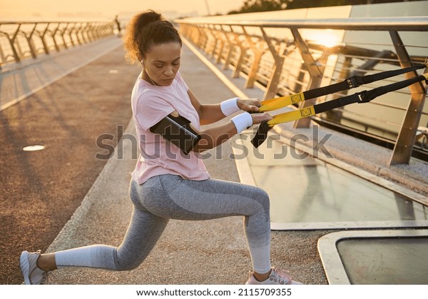 Athletic sportswoman exercises with suspension\
straps during body weight training at the urban environment of a\
city bridge. Full body female athlete in sportswear pulling\
suspension ropes and\
lunging