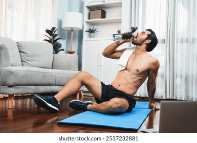 Athletic and sporty man drinking water on fitness mat after finishing home body workout exercise session for fit physique and healthy sport lifestyle at home. Gaiety home exercise workout training. - Powered by Shutterstock