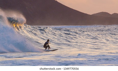 Athletic Surfer Dude Carving Large Breaking Wave At Colorful Summer Sunset. Reflective Ocean Waves Carrying Young Man On Surfboard To Tranquil Beach On A Beautiful Evening. Radiant Marine Landscape