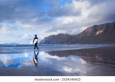 An athletic surfer man walks the beach with a surfboard. Sport travel destinaiton. Surfing lifestyle. Famara beach, Lanzarote, Canary Islands, Spain - Powered by Shutterstock