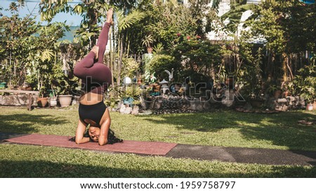 An athletic and tanned asian female yogi enters a headstand or Sirsana pose using a split-leg version. Practicing yoga outdoors at home on a sunny morning.