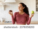 Athletic woman in activewear choosing apple fruit or donut struggling from sugar cravings during diet, standing in kitchen setting. Unhealthy vs healthy food choice concept