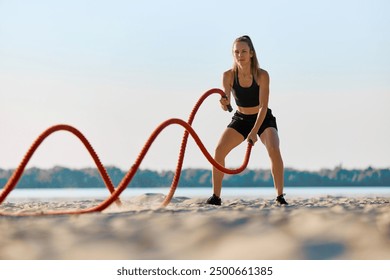 Athletic woman in black sportswear exercising with red battle ropes on a sandy beach, with water and trees in background under clear sky. Concept of sport, active and healthy lifestyle, endurance - Powered by Shutterstock