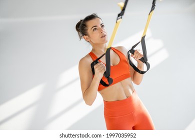 Athletic woman in bright sportswear doing cardio training on suspension straps at white gym. TRX training on fitness straps
