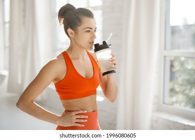 Athletic Woman In Bright Sportswear Drinking Protein Beverage From A Shaker, Standing Indoors. The Concept Of Sports Nutrition During Exercise