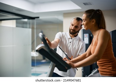 Athletic Woman Cycling On Exercise Bike While Personal Trainer Is Assisting Her During Sports Training At Health Club. Focus Is On A Coach. 