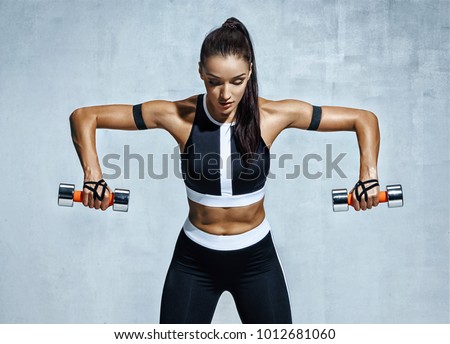 Athletic woman doing exercise for arms. Photo of muscular fitness model working out with dumbbells on grey background. Strength and motivation
