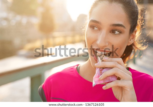 Athletic woman eating a\
protein bar. Closeup face of young sporty woman resting while\
biting a nutritive bar. Fitness beautiful woman eating a energy\
snack outdoor.