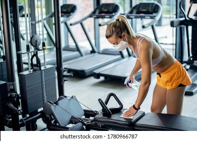 Athletic woman with face mask cleaning exercise machine with disinfectant while exercising in a gym during coronavirus epidemic. 