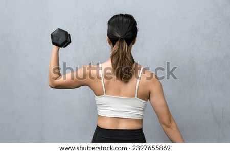 An athletic woman firmly grasps a hefty dumbbell behind her back, showcasing strength and determination. She stands in a well-lit gym atmosphere, the picture of healthful energy and dedication.