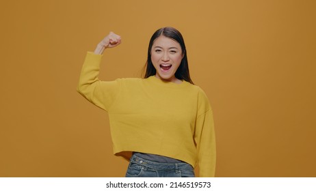 Athletic Woman Flexing Arms Muscles Over Yellow Background, Showing Muscular Body Before Workout Training. Confident Person Posing To Show Biceps, Clenching Fists. Self Assured Athlete