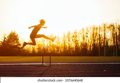 Athletic woman jumping above the hurdle on stadium running track during sunset - Shutterstock ID 1076640668