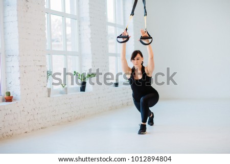 Athletic woman performing a functional exercise with suspension cable over white interior. 