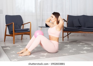 An Athletic Woman In Pink Sports Leggings And A Top With Her Hands Behind Her Head And A Ball Clenched Between Her Knees Does Twists In The Living Room Exterior.