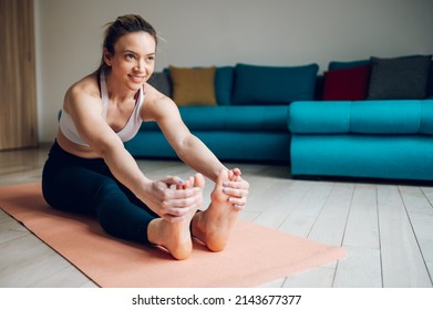 Athletic Woman Reaching Out To Touch Her Toes While Sitting On A Yoga Mat And Training At Home On The Floor. Stretching Before Exercising. Seated Forward Bend Pose.