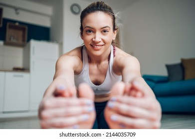 Athletic Woman Reaching Out To Touch Her Toes While Sitting On A Yoga Mat And Training At Home On The Floor. Stretching Before Exercising. Seated Forward Bend Pose.