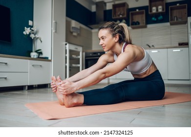 Athletic Woman Reaching Out To Touch Her Toes While Sitting On A Yoga Mat And Training At Home On The Floor. Stretching Before Exercising. Seated Forward Bend Pose.