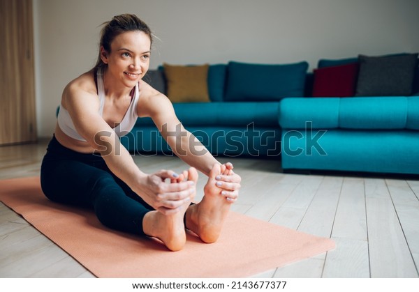 Athletic woman reaching out to\
touch her toes while sitting on a yoga mat and training at home on\
the floor. Stretching before exercising. Seated forward bend\
pose.