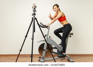 Athletic Woman Recording Tutorial Video On Cell Phone On Tripod, Riding Exercise Bike And Showing Thumb Up, Wearing Sports Tights And Red Top. Indoor Studio Shot On Gray Wall Background.