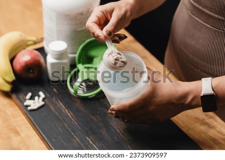 Athletic woman in sportswear with measuring spoon in her hand puts portion of whey protein powder into a shaker on wooden table with amino acid white capsules, bananas and apple, making protein drink.