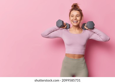 Athletic woman in sportswear raises dumbbells pumps biceps in gym being full of energy leads healthy lifestyle has happy expression isolated over pink background copy space for advertisement - Powered by Shutterstock