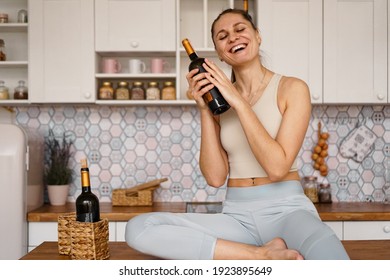Athletic woman in a tracksuit in a light kitchen drinks red wine from a bottle after doing sports. Woman laughs