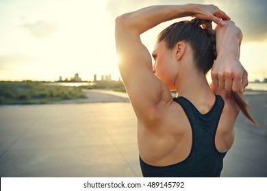 Athletic woman warming up before a workout standing facing the early morning rising sun on a rural road doing stretching exercises, close up with copy space