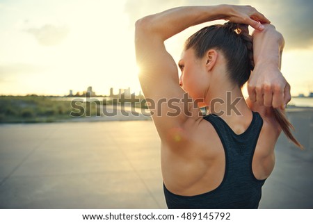 Athletic woman warming up before a workout standing facing the early morning rising sun on a rural road doing stretching exercises, close up with copy space