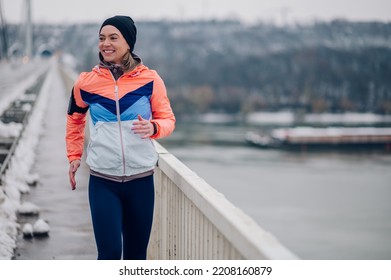 Athletic Woman Wearing An Active Wear And A Black Cap Jogging On A Bridge With Snow On Cold Winter Day. Winter Fitness. Copy Space. Fit Sportswoman Running In The City On Snow At Winter.