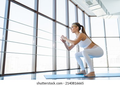 Athletic yoga woman in white sportswear doing squats barefoot on the mat indoors