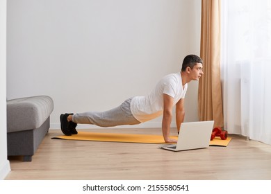 Athletic Young Adult Man In Sportswear Planking In Front Of Laptop, Having Online Fitness Class From Home In Living Room, Sporty Male Enjoying Daily Exercising.